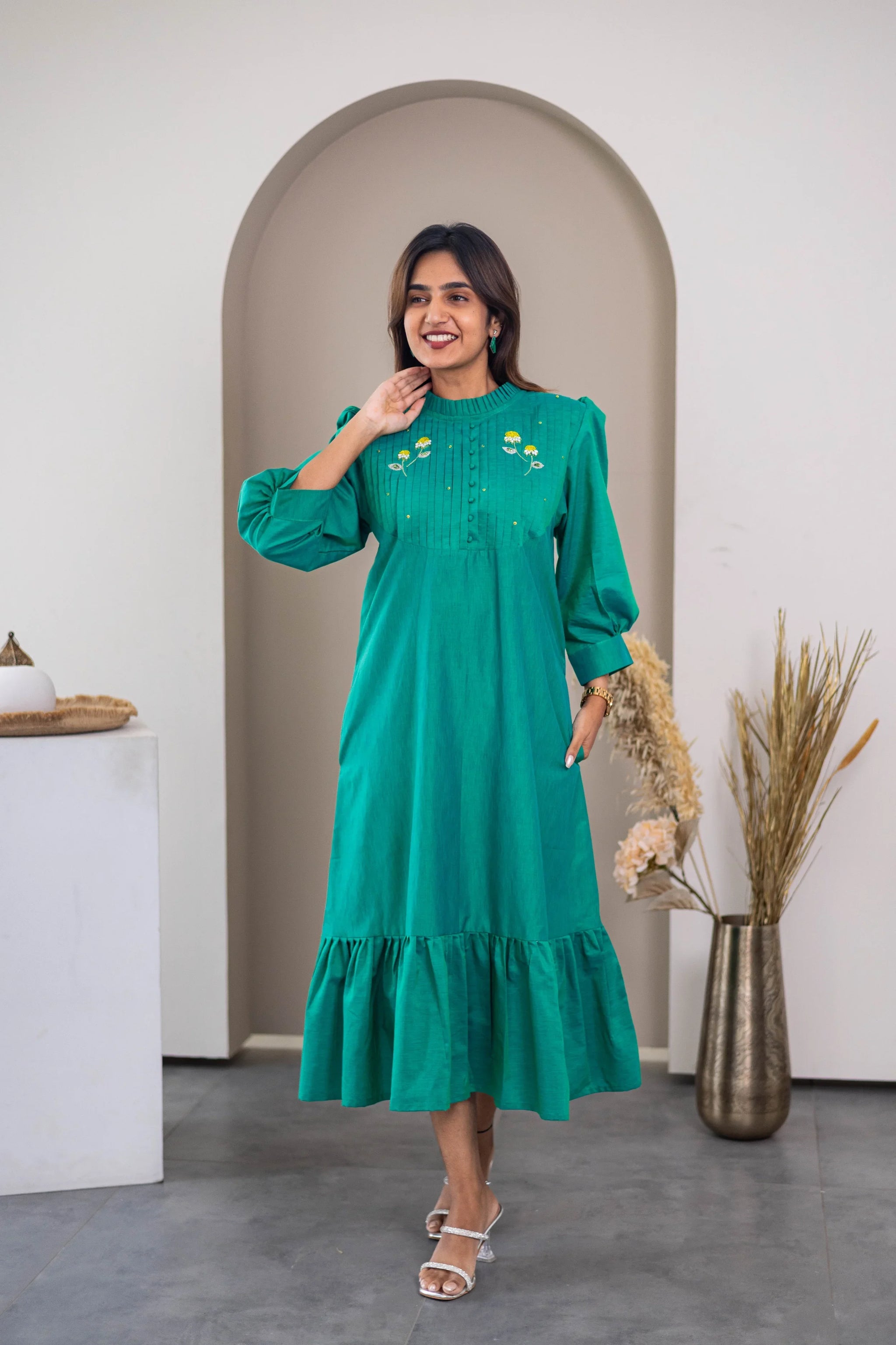 Woman wearing a green midi dress with floral embroidery and ruffle hem, standing indoors.