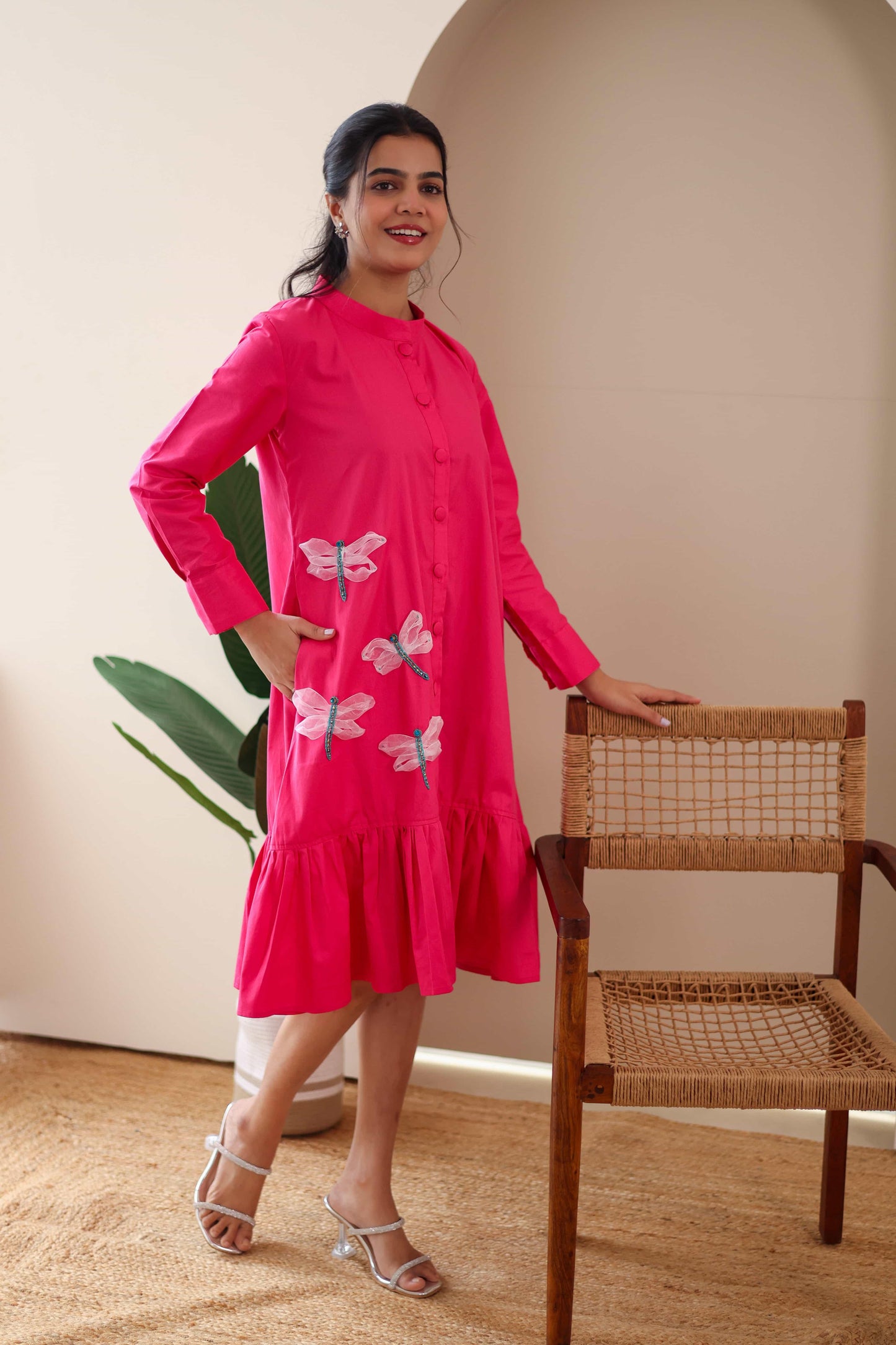 Woman in a bright pink cotton dress with dragonfly embroidery, standing indoors by a chair