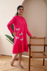 Woman in a bright pink cotton dress with dragonfly embroidery, standing indoors by a chair