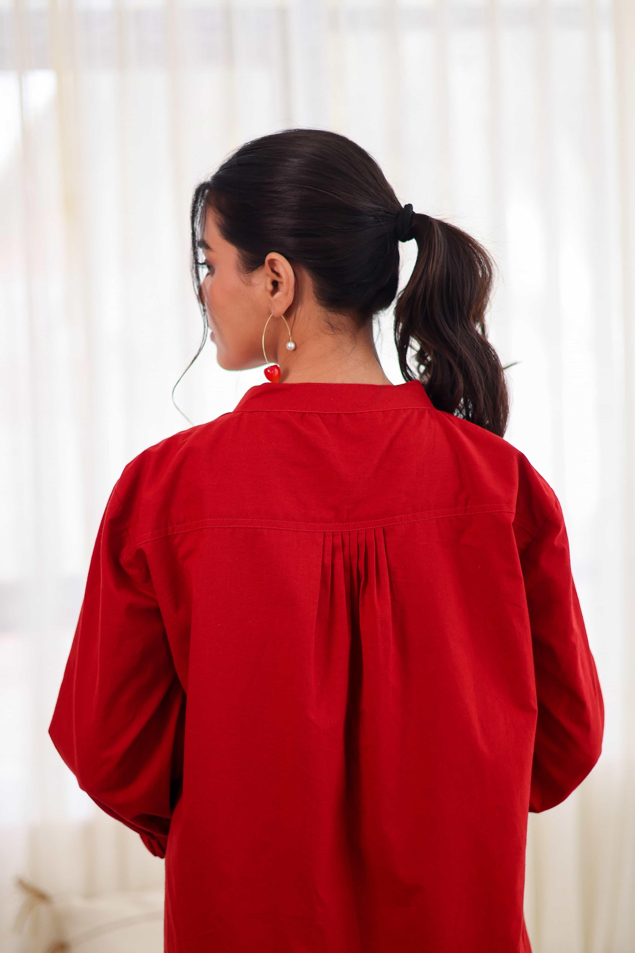 Woman wearing red cotton co-ord top with pleated back, gold hoop earrings
